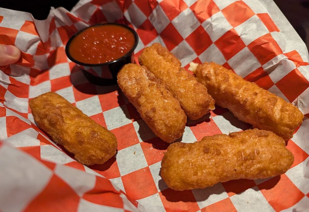 Cinco bastones de queso empanados y fritos en una cesta con papel de cuadros rojos, junto a un cuenco de salsa de tomate.