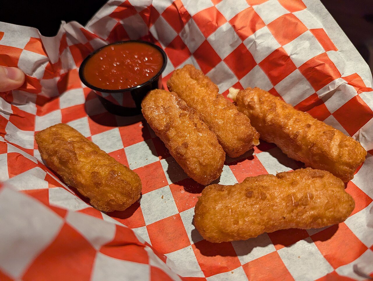 Cinco bastones de queso empanados y fritos en una cesta con papel de cuadros rojos, junto a un cuenco de salsa de tomate.