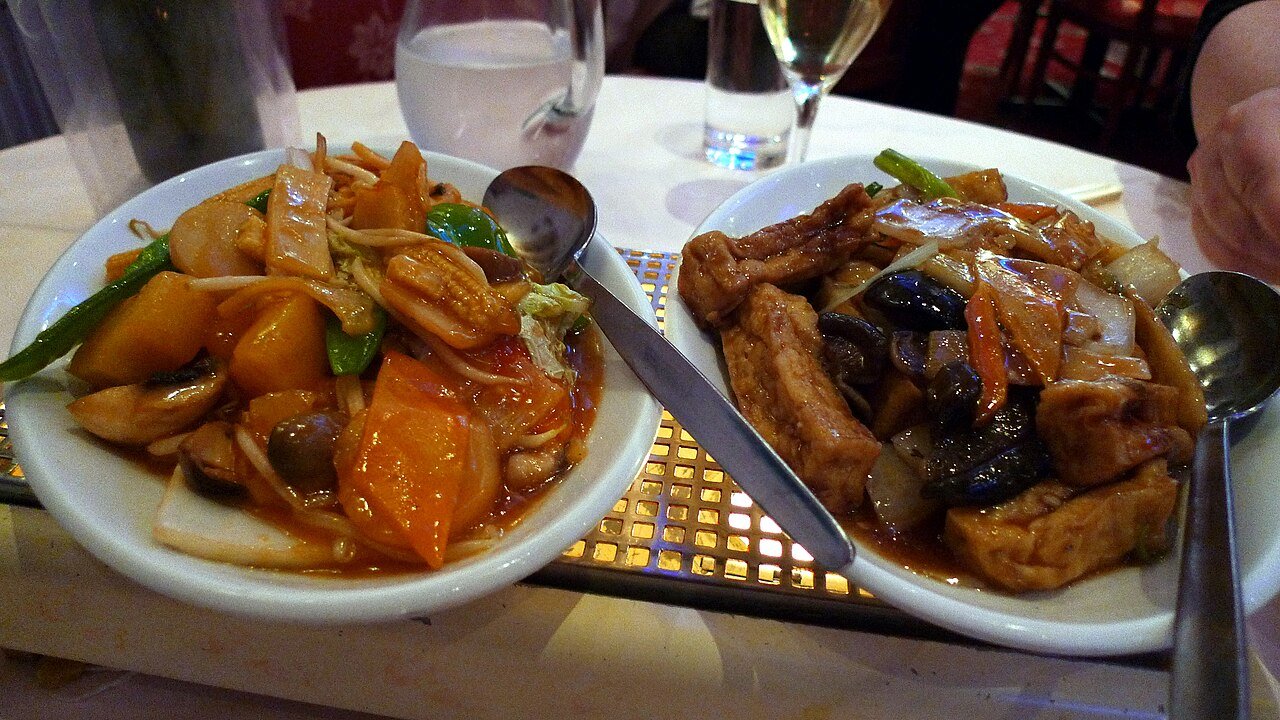 Dos cuencos blancos con platos asiáticos de verduras salteadas, tofu frito y salsa agridulce.