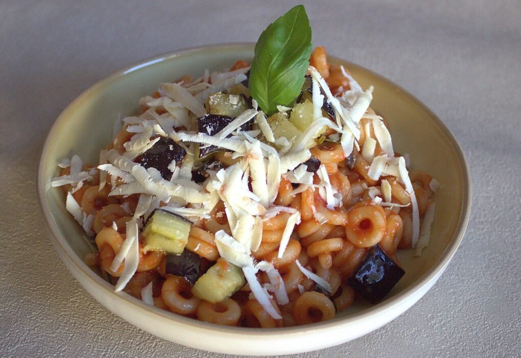 Un plato de pasta anular con salsa de tomate, cubos de berenjena, queso rallado y una hoja de albahaca.