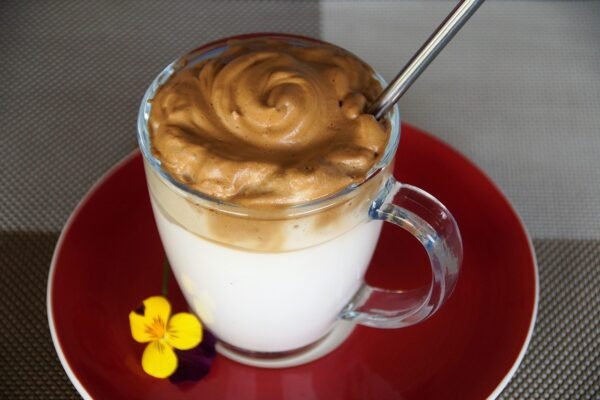 Un vaso de cristal con leche y una gruesa capa de crema de café dalgona encima, sobre un plato rojo.