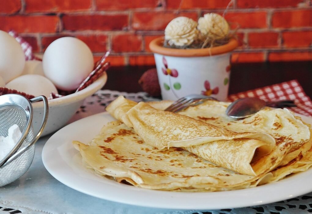 Una pila de crepes dorados en un plato blanco, junto a un bol de huevos y un colador con harina.