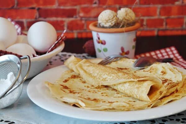 Una pila de crepes dorados en un plato blanco, junto a un bol de huevos y un colador con harina.