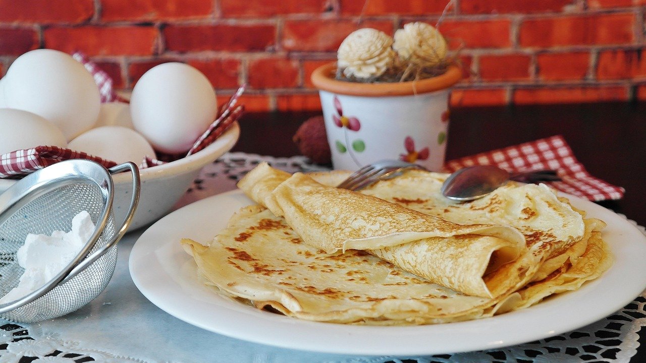 Una pila de crepes dorados en un plato blanco, junto a un bol de huevos y un colador con harina.